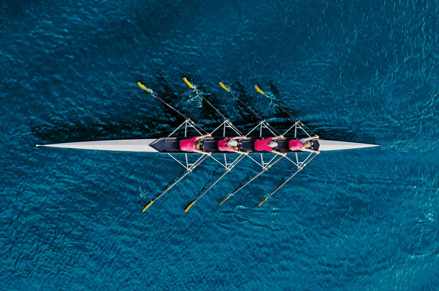 aerial image of a thin rowing boat with 4 teammates rowing in blue water